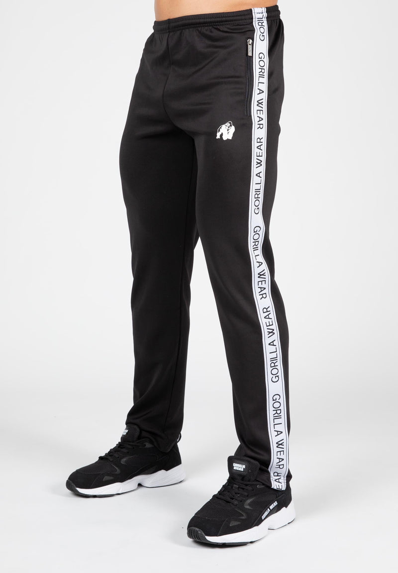 Load image into Gallery viewer, A person wearing black Delaware Track Pants with white taping on the sides and a logo on the thigh, paired with a white top and black shoes.
