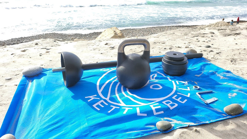 Load image into Gallery viewer, Kettlebell and dumbbells on a blue mat with 'Kettlebell' branding on a beach.